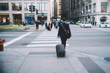 Back view of formally dressed banker walking on crossroad with baggage during business trip to megalopolis,young male entrepreneur with suitcase luggage stolling on urban setting of downtown .