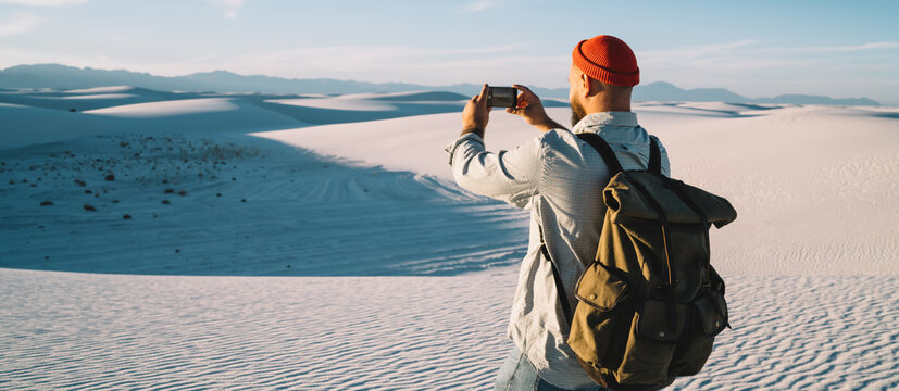 Man with touristic backpack using app on smartphone for making pictures in desert traveling on vacations, back view of hipster guy wanderlust taking photo of White sands national park on cellphone