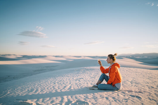 Tourist woman sitting on dunes with smartphone making picture of scenic view of desert at sunset, female tourist using telephone camera for taking photo of natural beauty of White sands scenery view