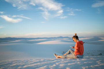 Young woman using smartphone sending pictures from trip satisfied with data connection in White Sands desert,hipster girl sitting on dune enjoying journey to New Mexico using mobile phone for chatting