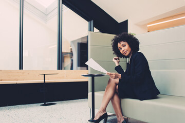 Portrait of attractive curly dark skinned businesswoman looking at camera sitting in modern designed office, charming prosperous 20s african american manager checking company finance reports