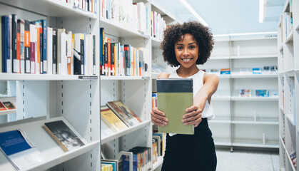 Cheerful 20s african american woman holding new book satisfied with new issue standing in bookstore looking in camera,portrait smiling prosperous dark skinned female librarian showing new book