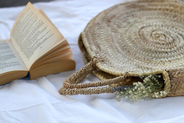 Wicker bag with flowers and open book on a picnic blanket. Selective focus.