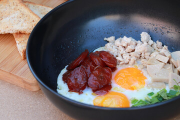 pan fried egg with toppings, Chinese sausage and pork sausage in the black pan with slice of bread.