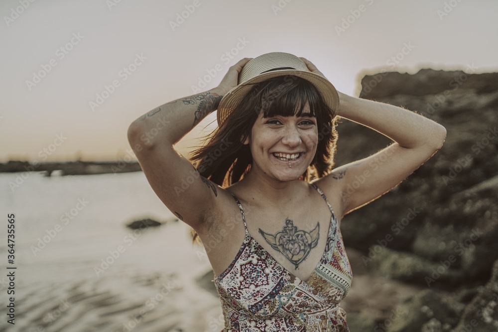 Sticker young female with tattoos wearing a dress and straw hat on blurred ocean background