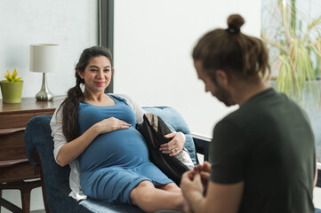 Beard man doing foot massage his pregnant wife while sitting on sofa in they home.