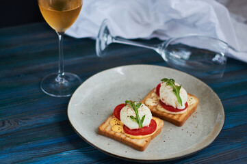 Large gray ceramic plate with toast and poached eggs, greens, tomatoes, sauce. Tasty breakfast. Glasses of wine.