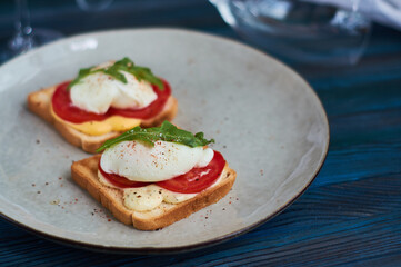 Large gray ceramic plate with toast and poached eggs, greens, tomatoes, sauce. Tasty breakfast. Glasses of wine.