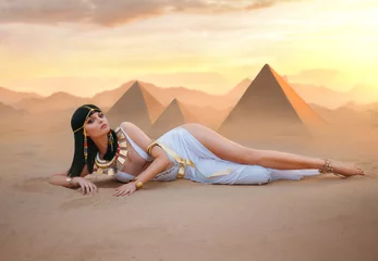 Reiche Luxusfrau im ägyptischen Stil. Sexy schöne Mädchengöttin Königin Cleopatra liegt auf gelben Sandwüstenpyramiden. Art altes Pharaonenkostüm weißes Kleid Goldzubehör schwarze Haarperücke ägyptisches Make-up © kharchenkoirina
