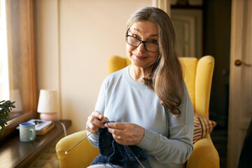 Retired people, aging, leisure, rest and hobby concept. Charming Caucasian grandmother in eyewear enjoying retirement, sitting at home in armchair, holding yarn and needles, knitting sweater