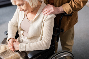 cropped view of senior man touching shoulder of disabled wife sitting in wheelchair with bowed head