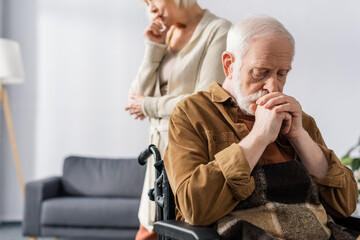 cropped view of sad senior woman standing near disabled, sick husband sitting in wheelchair with clenched hands