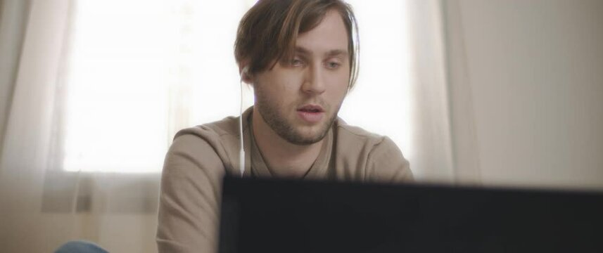 A young man speaking to someone through earphones while working on his laptop. Work from home concept. Slow motion, BMPCC 4K
