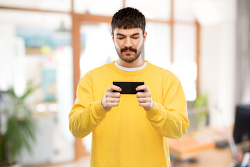 technology, communication and people concept - young man playing game on smartphone over office background