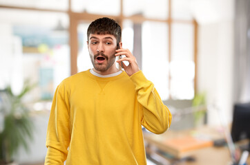 technology and people concept - shocked young man in yellow sweatshirt calling on smartphone over office background