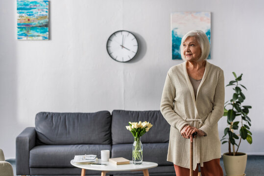 Smiling Senior Woman Looking Away While Standing With Walking Stick Near Bouquet Of Fresh Tulips On Coffee Table