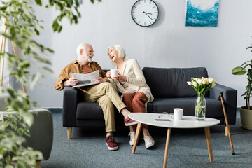 selective focus of happy senior couple laughing while talking on sofa near coffee table with tulips - Powered by Adobe
