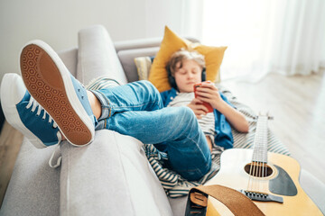 Preteen boy lying with guitar on cozy sofa dressed casual jeans and new sneakers listening to music and chatting using wireless headphones connected with a smartphone. Using electronic devices concept
