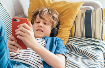 Preteen boy lying at home on cozy sofa dressed casual jeans and new sneakers listening to music and chatting using wireless headphones connected with smartphone. Child use electronic devices concept