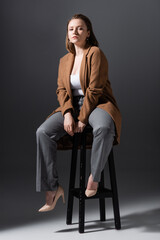 full length view of confident, pretty overweight girl sitting on high chair and looking at camera while posing on grey