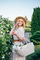 Portrait of Beautiful caucasian girl with long hair and straw hat in dress. Walking, smiling in summer park in blossom bush of white roses flowers 