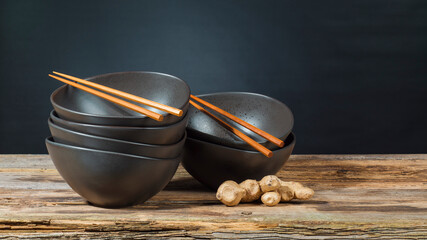 Two stacks of empty dark brown rice bowls with two pairs of wooden chopsticks and a ginger tuber stand on an old wooden table in front of a black background. - Powered by Adobe