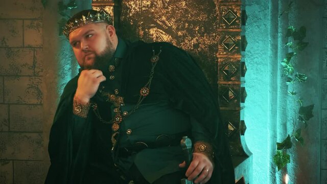 warlike medieval courageous strong man sits on old bronze throne, image tyrannical king. Carnival vintage costume velvet clothes Royal crown, jewelry gems. Serious pensive face. gothic castle room 