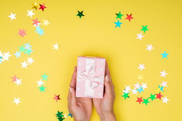 Women hands holding a gift or gift box decorated with confetti on a yellow background top view.
