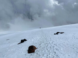 A climber with a backpack and trekking poles walks along a snowy path.
