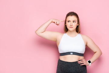 thoughtful overweight girl pointing with finger at her head on pink