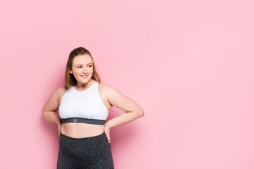 Fototapeta na wymiar cheerful overweight girl in sportswear holding hands on hips and looking away on pink