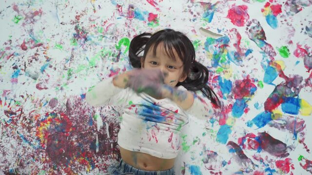 Top view of adorable smiling asian little girl is laying down and playing her painted hands on her abstract artwork at home, concept of freedom, free play and art education for kid learning at home.
