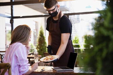 The waiter works in a restaurant in a medical mask, gloves during coronavirus pandemic. Representing new normal of service and safety. Giving dessert, tasty fruit salad. Taking care of clients.