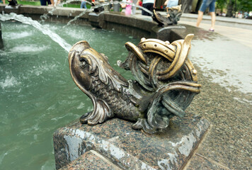 Metal fish on the fountain near the pedestrian-bicycle bridge. Splashing water from the fish's mouth. Bronze fish fountain.