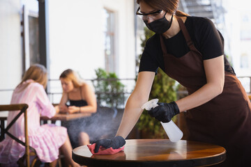 The waitress works in a restaurant in a medical mask, gloves during coronavirus pandemic. Representing new normal of service and safety. Disinfecting table, surfaces. Taking care of clients.