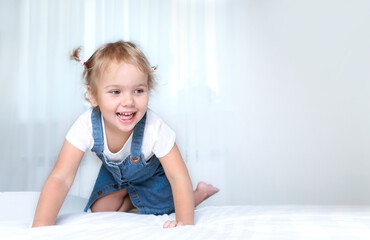 Fototapeta premium Child on a bed empty copy space background.Girl in white sheet in bedroom.Happy kid jumping on bed.
