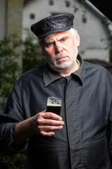 Serious kind sad elderly man with black old-fashioned old clothes and a leather cap with a glass of beer in his hand. Looks at the camera. Vertical orientation.