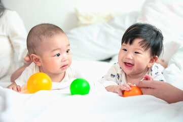 Obraz na płótnie Canvas Two cute little Asian babies boy lying on the white bed and playing with colorful ball. Baby playing together.