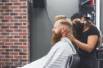 Naklejka premium Female barber in mask cuts a man hairs with hair clipper. Hairstyle during social distancing