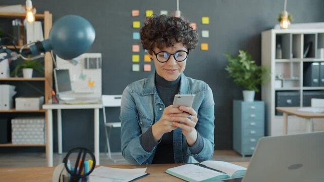 Beautiful woman is using smartphone touching screen smiling sitting at desk in office room alone enjoying gadget. People, workspace and devices concept.