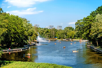 city park landscape at daytime against blue sky with clouds background. People on pleasure boat on sunny day in pond at city park. Summer time in Moscow city Russia