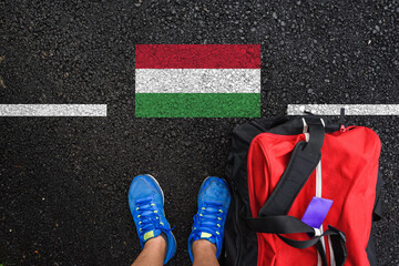 a man with a shoes and travel bag is standing on asphalt next to flag of Hungary and border
