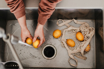 Yellow lemons are in net cotton shopping bag in sink. Washing fruits before using them. Local farm produce. Fresh seasonal food. Woman washes fruit. Zero waste, healthy. From farm to table. Citrus