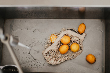 Yellow lemons are in net cotton shopping bag in sink. Washing fruits before using them. Local farm produce. Fresh seasonal food. Bright color. Zero waste, organic, healthy. From farm to table. Citrus