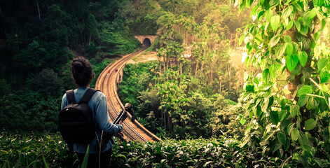 Young woman backpacker photographer looks at the Demodara nine arches bridge from tea and pepper plantation in the morning sunlight, the most visited sight of Ella town in Sri Lanka.