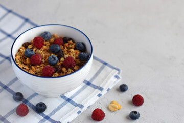 Breakfast or a light snack of muesli, oatmeal and fresh raspberries and blueberries in a white plate with a blue border on a light concrete background with space for text.