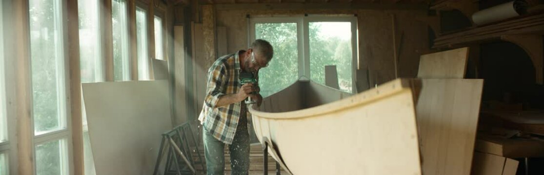 Mid 50s Caucasian male building a canoe in his workshop, using milling tool. Boat making hobby, small business owner. Shot on RED Dragon