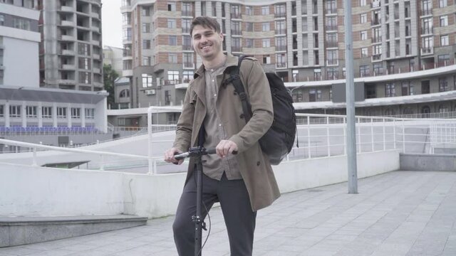 Portrait of cheerful adult Caucasian man standing on city street with scooter and smiling at camera. Young brunette guy having fun outdoors. Individuality, lifestyle, happiness.