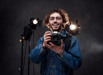 Young caucasian man in glasses wearing denim jacket holds a digital camera, laughs and looking on camera. Dark photo studio with lighting equipment in the background