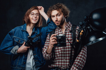 The team of two young photographers holds a digital camera and lighting equipment posing in studio